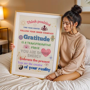 Könnte beinhalten: Ein gerahmter Kunstdruck mit einem goldenen Rahmen mit den Worten "Think positive" und "Gratitude" in verschiedenen Farben. Der Druck enthält inspirierende Sätze und Symbole wie eine Lotusblume, ein Herz und einen Schmetterling. Die Kunst wird von einer Person gehalten.