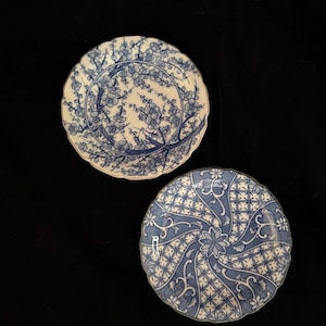 May include: Two round ceramic plates with scalloped edges. The top plate features a blue and white floral branch pattern. The bottom plate displays a blue and white geometric grid design with swirling vine motifs and a central flower element.