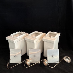 May include: Three white rectangular ceramic canisters with open tops, accompanied by three square metallic lids and three matching metal ladles with curved handles, all arranged in a row against a dark background.