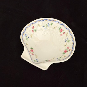 May include: A white porcelain shell-shaped dish featuring a delicate floral pattern with small pink and blue flowers. The rim is decorated with a blue and gold leaf-style border, and the interior surface has a subtle embossed texture.
