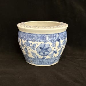 May include: A white ceramic planter featuring a blue hand-painted floral pattern and a geometric Greek key border around the rim. The bowl-shaped vessel has a wide opening and a tapered base, decorated with repeating botanical motifs in blue glaze.