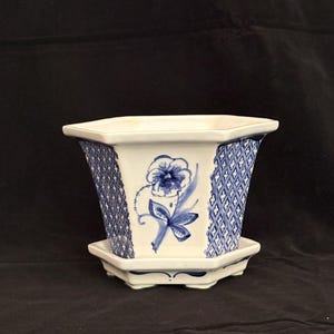 May include: A hexagonal ceramic planter with a matching saucer, featuring a white base with blue hand-painted floral motifs on the front panels and a blue and white lattice pattern on the side panels, finished with a glossy glaze and small feet.