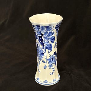 May include: A white ceramic vase with a flared hexagonal rim and base, featuring a blue floral pattern painted across the exterior. The design includes detailed blossoms and leaves in varying shades of blue, with a smooth, glossy finish throughout.