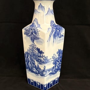 May include: A hexagonal ceramic vase featuring a white base with blue hand-painted landscape illustrations of mountains, trees, and figures. The neck is cylindrical with a flared rim, and the base is decorated with a repeating blue geometric pattern.