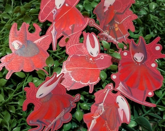Puede incluir: Una colección de siete pegatinas que representan al personaje Hornet del videojuego Hollow Knight. Cada pegatina muestra al personaje con una capa roja, una máscara blanca y un arma de aguja, en diversas poses dinámicas y estilos artísticos.