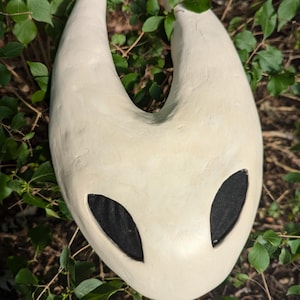 Puede incluir: Una máscara de cerámica blanca con agujeros negros para los ojos, que se asemeja a un personaje de un videojuego. La máscara tiene forma de media luna con bordes redondeados.