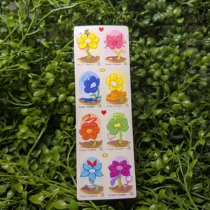 May include: A rectangular sticker sheet featuring eight distinct cartoon flower illustrations arranged in two columns. Each flower is enclosed in a rounded arch frame with the text Flower Kingdom printed below. The flowers are yellow, pink, blue, orange, red, green, and purple.