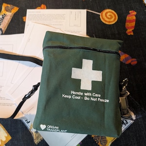 May include: A dark green rectangular bag with a black adjustable strap, featuring a white cross symbol, the text "Handle with Care Keep Cool - Do Not Freeze," and "ORGAN TRANSPLANT" with a heart icon. Several white paper instruction sheets are included.