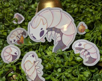 Puede incluir: Una colección de pegatinas con una criatura insectoide estilizada, blanca y púrpura, con cuerpo segmentado y ojos oscuros. Las pegatinas están dispuestas sobre un lecho de follaje verde, con pequeñas campanas doradas.