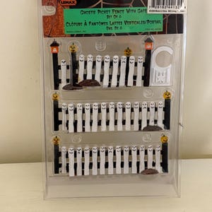 May include: A set of six Ghostie Picket Fences with gates. Each fence is white with ghost faces and is accented with black posts and orange pumpkin lights. The fences are packaged in a clear plastic container.