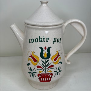 May include: White ceramic cookie jar shaped like a teapot with a green lid and handle. The jar has a green and white floral design with red and yellow accents. The text "cookie pot" is written in green on the front of the jar.