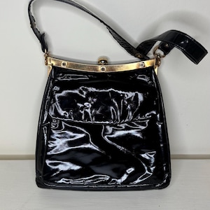May include: A black patent leather handbag with a glossy finish, featuring a gold-tone metal frame closure at the top and a single matching black patent leather shoulder strap. The bag has a structured rectangular shape with a smooth, reflective surface.