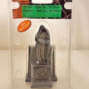 May include: A Lemax Spooky Town Collection figurine titled Crypt - Rise All Ye Dead, featuring a grey stone-colored statue of a cloaked figure standing on a tombstone base with the text Rise All Ye Dead, encased in original clear plastic packaging.