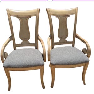May include: Two armchairs featuring light beige wooden frames with a distressed finish, curved armrests, and open-back designs with a central vertical splat. The seats are upholstered in a textured gray fabric, supported by four curved legs.