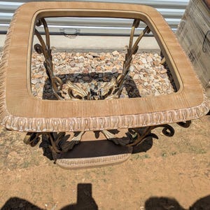 May include: Rectangular coffee table with a light wood-tone frame and ornate gold-colored metal supports. The table features a decorative edge and a lower shelf. The design is vintage and elegant.