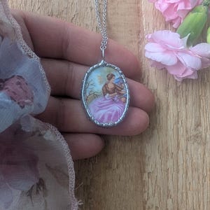 May include: Silver necklace with an oval pendant. The pendant displays a miniature painting of a woman in a pink dress, framed by a decorative silver border. Pink flowers and a wooden surface are in the background.