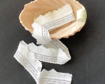 May include: A length of white cotton lace trim with a scalloped edge and a central band of embroidered floral eyelet details, coiled inside a pale peach-colored ceramic dish shaped like a shell with a smooth, glossy finish and curved edges.