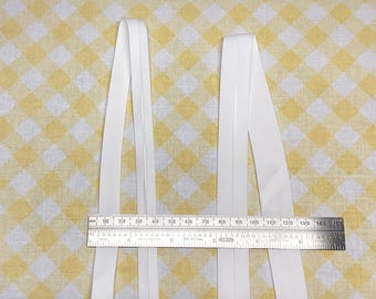 May include: Two strips of white bias tape are positioned vertically behind a metal ruler. The ruler shows the width of the tape strips is approximately 0.5 inches each, with a total span of about 5.5 inches across the two pieces of fabric.