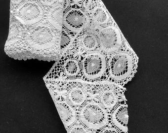 May include: A roll of white lace trim featuring a repeating pattern of circular motifs and small floral accents. The intricate needlework displays a delicate openwork design with scalloped edges, showcasing a continuous decorative textile strip.