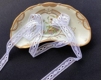 May include: A length of white lace trim with a repeating oval pattern resting on a semi-circular ceramic dish. The dish features a cream-colored center with a painted illustration of a figure in a garden, surrounded by a gold-trimmed scalloped edge.
