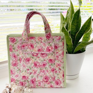 May include: A floral tote bag with a pink rose pattern on a quilted fabric. The bag has a green border and handles, and a front pocket. The bag is sitting next to a potted plant and seashells.
