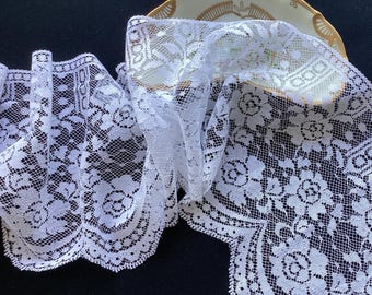 May include: A length of white lace trim featuring a detailed floral rose pattern with a scalloped edge. The delicate fabric is draped over a small, cream-colored ceramic dish with a gold-painted rim, showcasing the intricate mesh and weave.