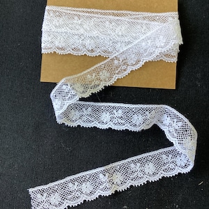 May include: A length of white lace trim featuring a delicate floral pattern and scalloped edges, wrapped around a rectangular piece of brown cardboard. The lace has a fine mesh texture and is displayed in a loose, winding S-shaped configuration.