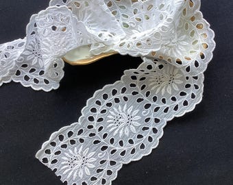 May include: A white eyelet lace trim featuring a repeating floral pattern with circular cutouts and scalloped edges. The fabric is draped in a continuous length, showcasing intricate embroidery and openwork details throughout the entire design.