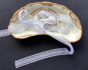 May include: A length of white lace trim with a repeating oval and dot pattern coiled inside a crescent-shaped ceramic dish. The dish features a cream-colored base, a gold-painted scalloped rim, and a central floral design with green leaves.