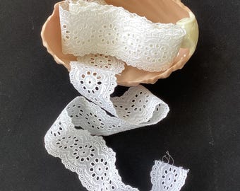May include: A length of white eyelet lace trim featuring a repeating floral pattern with circular cutouts and scalloped edges, coiled within a pale peach-colored scalloped dish. The lace displays intricate embroidery and a delicate openwork design.