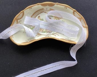 May include: A length of white cotton ribbon with a delicate hemstitch border, coiled and draped over a crescent-shaped porcelain dish featuring a gold-trimmed edge and a central floral illustration with a small figure in a garden setting.