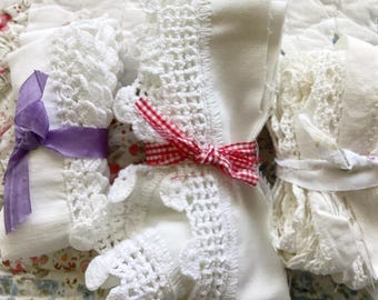 May include: Three rolls of white fabric trim featuring intricate crochet lace edges. The left roll is tied with a purple satin ribbon, the center roll is secured with a red and white gingham ribbon, and the right roll is tied with a plain white fabric strip.