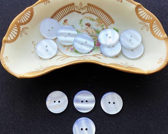 May include: A collection of round, two-hole buttons with a smooth, iridescent light blue finish. Several buttons are arranged inside a curved, cream-colored ceramic dish with gold-painted edges and a central floral illustration, while four buttons lie below.