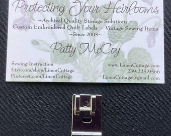 May include: A silver-colored metal sewing machine presser foot with a rectangular base and a central attachment bar, positioned below a white business card featuring floral illustrations and the text Protecting Your Heirlooms, Patty McCoy.