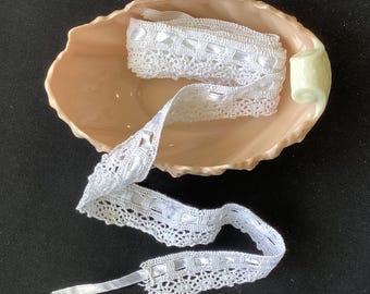 May include: A length of white lace trim featuring a woven ribbon detail, coiled inside a pale peach-colored ceramic dish with a scalloped edge and a small handle on one side. The trim has a delicate, openwork pattern along the bottom edge.