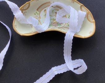 May include: A length of white cotton trim with a scalloped edge and a central row of eyelet detailing, coiled over a crescent-shaped ceramic dish featuring a painted scene of figures and gold-toned decorative trim along the scalloped edges.