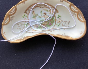 May include: A crescent-shaped porcelain dish with a cream-colored base and gold-painted scalloped edges. The center features a painted scene of two figures in a garden setting. A thin white elastic cord is draped across the surface of the dish.