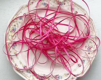 May include: A tangle of thin, vibrant hot pink satin ribbon rests inside a vintage-style ceramic plate. The plate features a scalloped edge with a delicate, repeating floral pattern in soft pastel shades of pink, blue, and green on a cream background.