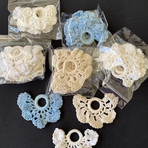 May include: A collection of handmade crochet doilies in various colors, including white, cream, and blue. Some are packaged in clear plastic bags. The doilies have a floral design with a central ring.