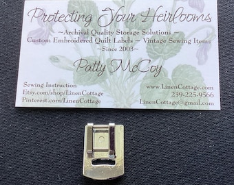 May include: A silver-colored metal sewing machine presser foot with a rectangular base and a central attachment bar. Above the presser foot is a white business card with floral illustrations and text including Protecting Your Heirlooms.
