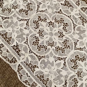 May include: An off-white lace table runner featuring an intricate repeating floral pattern with openwork mesh sections and solid embroidered edges. The textile displays a symmetrical arrangement of blossoms and scrolling vine motifs throughout.
