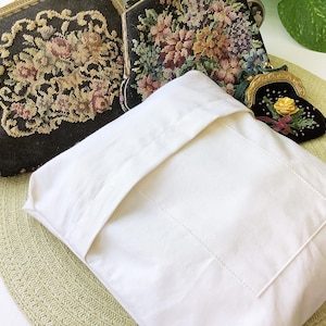 May include: A white pillowcase with a stitched pocket, resting on a light green woven mat. Two vintage tapestry purses with floral designs and gold frames are visible, along with a small black purse with a yellow rose.