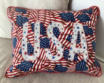 May include: Rectangular pillow with a patriotic design. The pillow features a pattern of American flags in red, white, and blue. The word "USA" is prominently displayed in white letters with blue and red stars. The pillow has a red border.