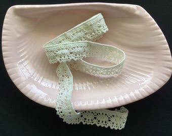 May include: A coiled length of pale cream-colored lace trim featuring a repeating floral pattern with scalloped edges. The delicate fabric strip is resting inside a smooth, light pink ceramic dish shaped like a large, ribbed seashell.