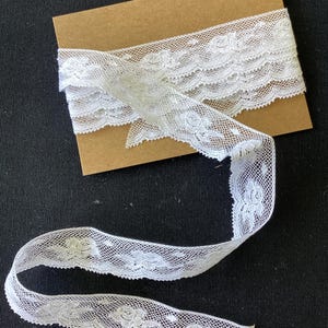 May include: A length of white floral lace trim wrapped around a rectangular brown cardboard card. The lace features a repeating rose pattern with a scalloped edge and a fine mesh background, with a portion of the trim unspooled and extending outward.