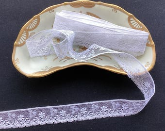 May include: A roll of white lace trim featuring a delicate floral pattern and scalloped edges, resting inside a crescent-shaped ceramic dish with a cream-colored base, gold-painted decorative trim, and subtle floral accents on the surface.