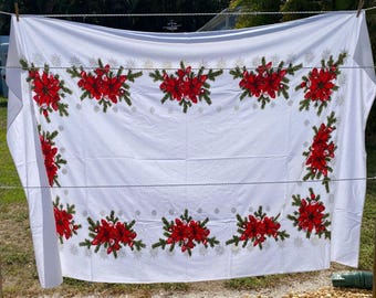 May include: A rectangular white tablecloth featuring a repeating border pattern of vibrant red poinsettia flowers, green pine branches, and small silver starburst accents arranged in clusters along the edges of the fabric.