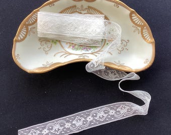 May include: A white lace ribbon with a repeating floral pattern and scalloped edges, coiled and draped over a crescent-shaped porcelain dish featuring a cream-colored base, gold-painted trim, and a central floral motif with green and pink accents.