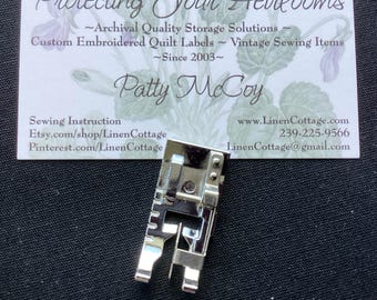 May include: A silver-colored metal sewing machine presser foot with a rectangular shank and a two-pronged base. Behind the presser foot is a white business card with floral illustrations and text including Protecting Your Heirlooms and Patty McCoy.