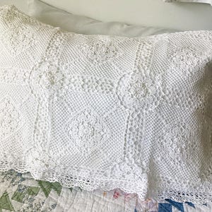 May include: A rectangular white pillow sham featuring an intricate crochet pattern composed of repeating square motifs with central floral designs, connected by a fine mesh grid, finished with a delicate scalloped lace border along the bottom edge.